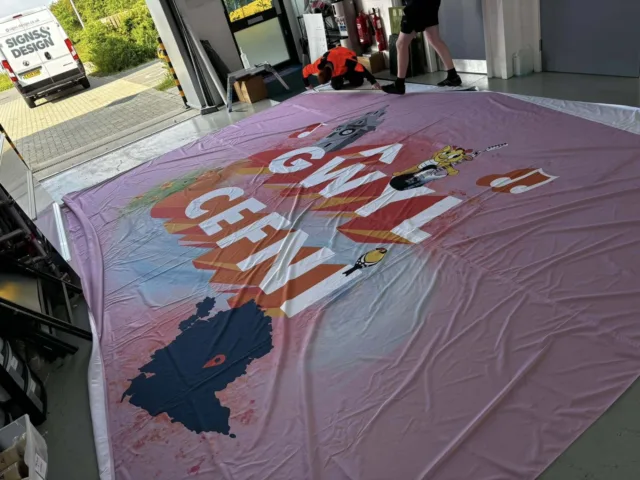 Oversized pink fabric event banner with colourful graphics and Welsh text, laid out on the workshop floor